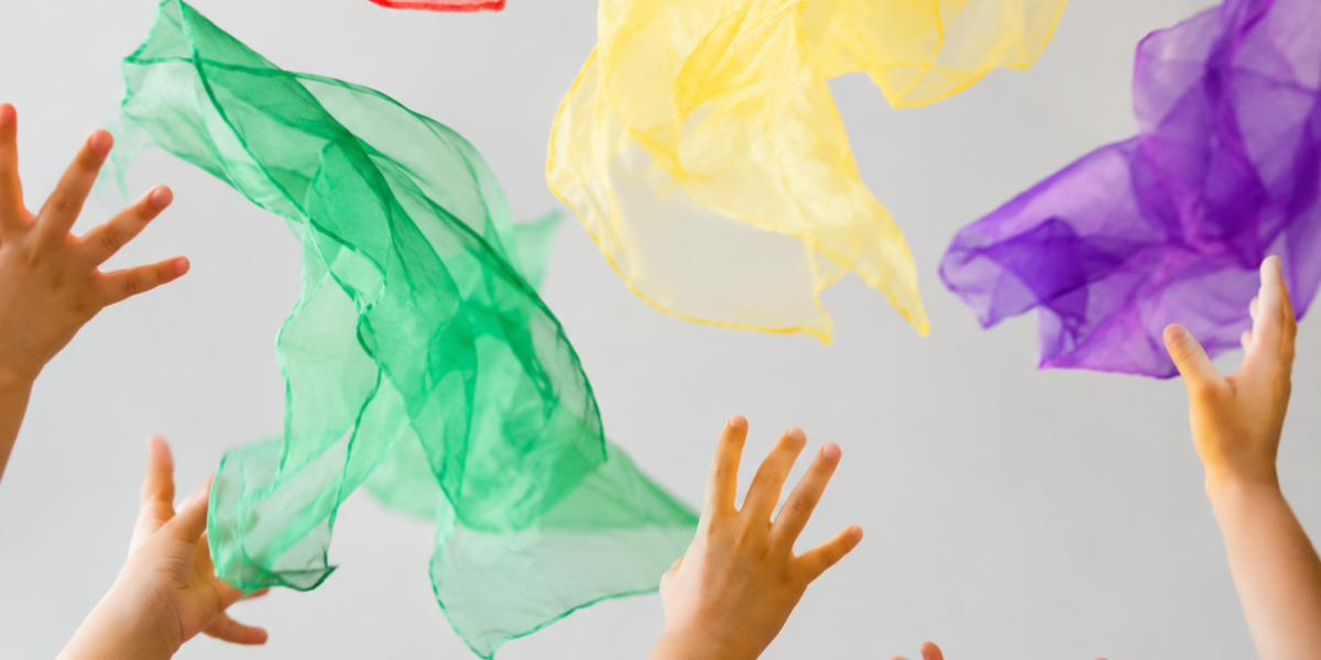 Verschiedene Kinderhände greifen nach bunten, transparenten Tüchern, die in der Luft schweben. Die Tücher sind in den Farben Rot, Blau, Gelb, Grün und Lila. Der Hintergrund ist hell und neutral.