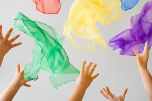 Verschiedene Kinderhände greifen nach bunten, transparenten Tüchern, die in der Luft schweben. Die Tücher sind in den Farben Rot, Blau, Gelb, Grün und Lila. Der Hintergrund ist hell und neutral.