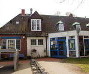 Ein Gebäude mit rotem Backstein und einem Satteldach, das mehrere Fenster hat. Der Eingangsbereich ist verglast und mit blauen Rahmen gestaltet. Vor dem Gebäude stehen einige Fahrräder, und im Garten sind kleine Blumenbeete zu sehen.
