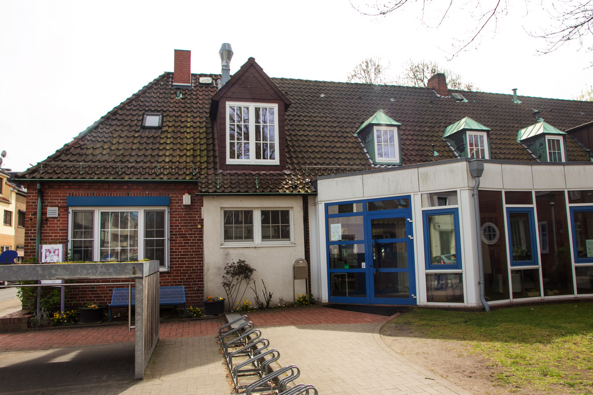 Ein Gebäude mit rotem Backstein und einem Satteldach, das mehrere Fenster hat. Der Eingangsbereich ist verglast und mit blauen Rahmen gestaltet. Vor dem Gebäude stehen einige Fahrräder, und im Garten sind kleine Blumenbeete zu sehen.