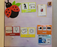 Das Bild zeigt ein buntes Wandbrett mit verschiedenen Symbolen und Illustrationen. Es enthält einem Marienkäfer, Karten mit einem Pinguin, einem Eisbären, einem Krokodil, einem Eis und zwei Herzen. Ein Abschnitt ist für den Mittwoch (Mi) gekennzeichnet, und es gibt eine freundliche Aufforderung zum Abschied.