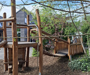 Ein Spielplatz mit einer Holzkonstruktion, die eine Rutsche und eine Brücke beinhaltet. Umgeben von Bäumen und Sträuchern, vermittelt die Szene eine einladende, natürliche Umgebung für Kinder zum Spielen und Entdecken.