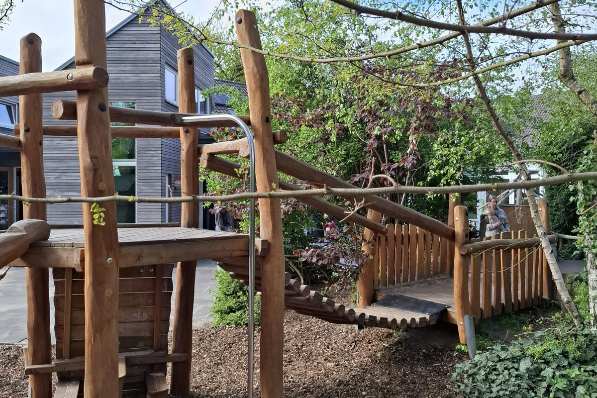 Ein Spielplatz mit einer Holzkonstruktion, die eine Rutsche und eine Brücke beinhaltet. Umgeben von Bäumen und Sträuchern, vermittelt die Szene eine einladende, natürliche Umgebung für Kinder zum Spielen und Entdecken.