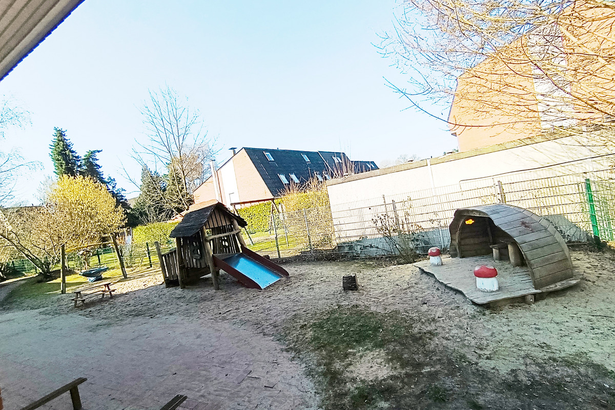 Spielplatz mit einem Holzspielhaus und einer Rutsche im Sandbereich. Daneben stehen zwei kleine Sitze und ein Tisch. Im Hintergrund stehen Bäume und eine Mauer, während darüber ein klarer blauer Himmel sichtbar ist.