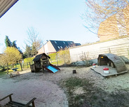 Spielplatz mit einem Holzspielhaus und einer Rutsche im Sandbereich. Daneben stehen zwei kleine Sitze und ein Tisch. Im Hintergrund stehen Bäume und eine Mauer, während darüber ein klarer blauer Himmel sichtbar ist.