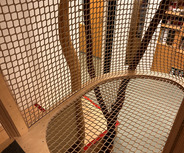 Ein Spielbereich aus Holz mit einem netzartigen Boden, der über einer Öffnung schwebt. Sichtbare Holzpfosten formen eine natürliche Struktur. Im Hintergrund ist eine Tür und einige Möbel zu erkennen.