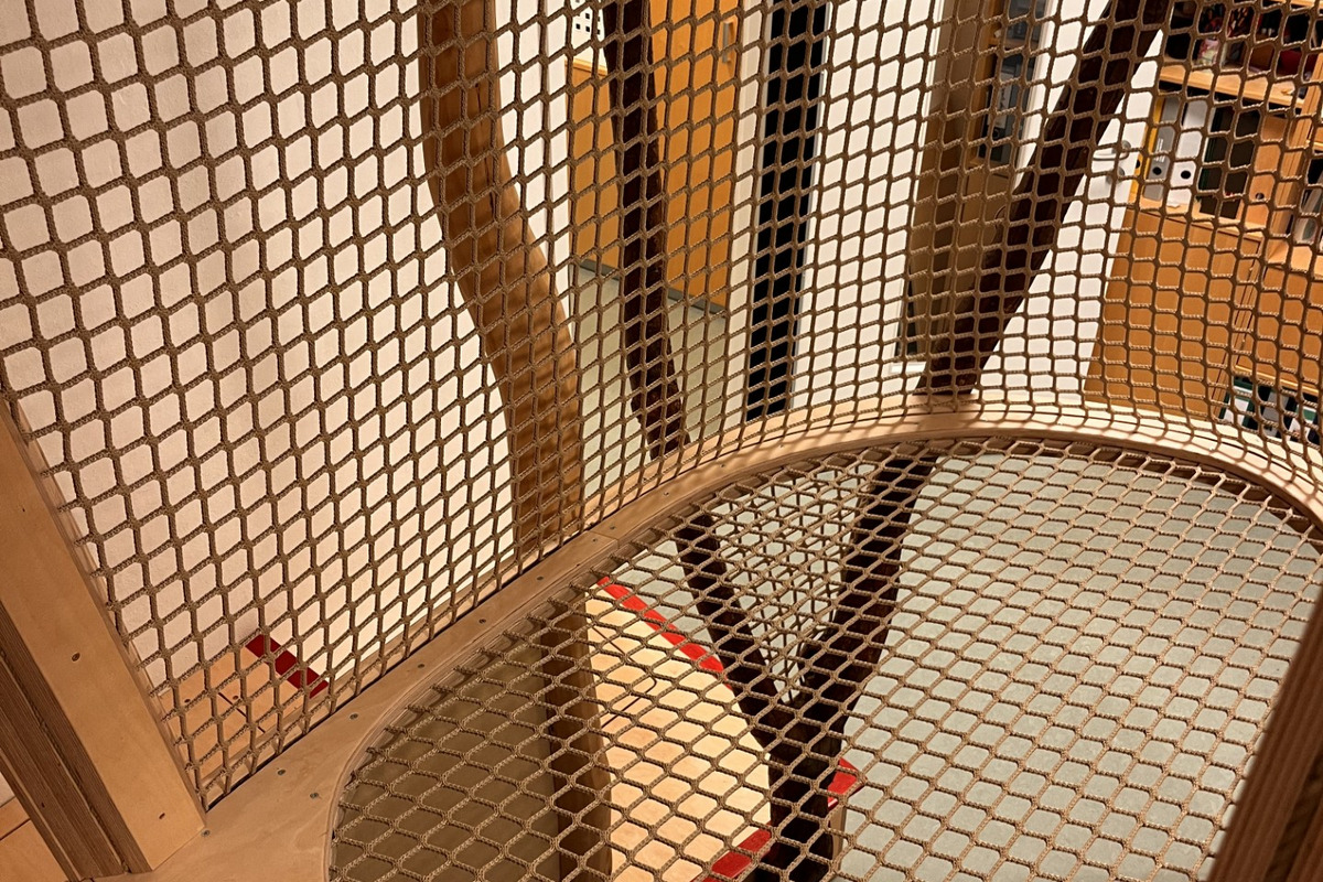 Ein Spielbereich aus Holz mit einem netzartigen Boden, der über einer Öffnung schwebt. Sichtbare Holzpfosten formen eine natürliche Struktur. Im Hintergrund ist eine Tür und einige Möbel zu erkennen.