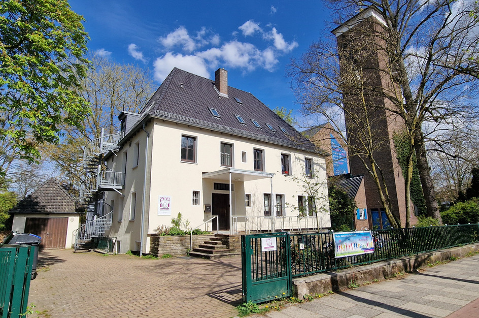 Ein dreigeschossiges, helles Gebäude mit einem Satteldach steht an einer Straße. Auf der linken Seite ist eine gewundene Feuerwehrleiter zu sehen. Im Hintergrund erhebt sich ein höherer, rechteckiger Turm. Umgeben von Bäumen und einer grünen Hecke ist der Eingangsbereich klar erkennbar.