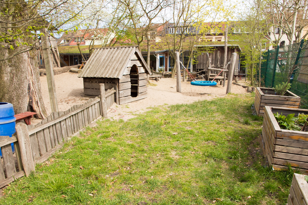 Ein Kinderspielplatz mit einem Holzspielhaus, einer Schaukel und einem Sandbereich. Bäume und ein Zaun umgeben den Spielplatz. Im Hintergrund sind einige Gebäude sichtbar. Der Boden ist durchgehend mit Gras und Sand bedeckt.