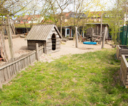 Ein Kinderspielplatz mit einem Holzspielhaus, einer Schaukel und einem Sandbereich. Bäume und ein Zaun umgeben den Spielplatz. Im Hintergrund sind einige Gebäude sichtbar. Der Boden ist durchgehend mit Gras und Sand bedeckt.