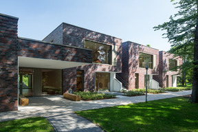 Moderne Gebäude aus roten Ziegeln, die in einem ruhigen Umfeld stehen. Große Fenster lassen viel Licht herein. Vor den Gebäuden verläuft ein gepflasterter Weg, umgeben von Rasen und einigen Büschen. Die Architektur wirkt klar und zeitgemäß.