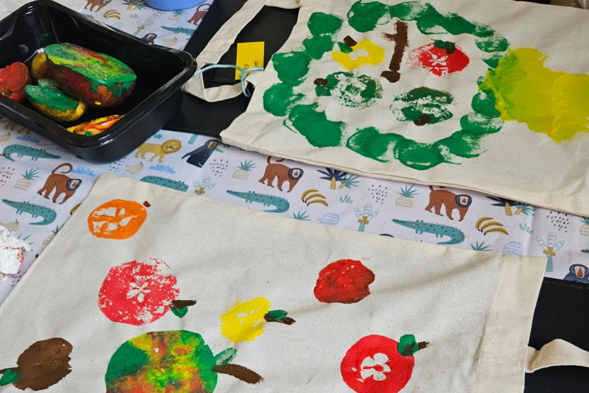 Zwei bemalte Stoffbeutel liegen auf einem Tisch. Der obere Beutel zeigt eine grüne Baummotivanordnung, während der untere Beutel bunte Obstmuster in Form von Äpfeln und anderen Früchten zeigt. Der Tisch ist mit einem Tiermuster überzogen, und einige Farbbehälter sind sichtbar.