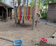 Auf dem Bild ist ein Spielplatz zu sehen. Im Vordergrund steht eine blaue Transportkiste, während im Hintergrund ein bunter Spieltunnel aus Holzpfählen und farbigen Brettern zu erkennen ist. Umgeben ist der Bereich von Sand und Bäumen.