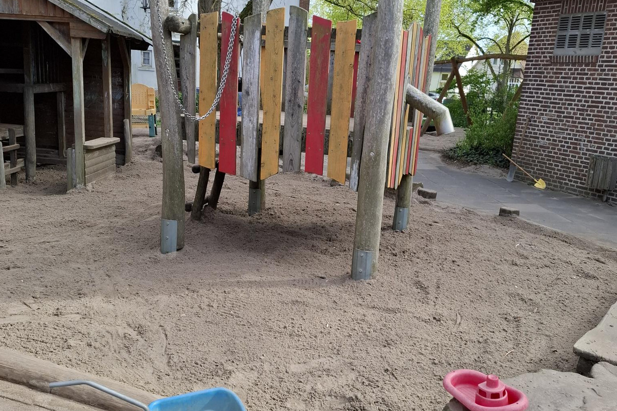 Auf dem Bild ist ein Spielplatz zu sehen. Im Vordergrund steht eine blaue Transportkiste, während im Hintergrund ein bunter Spieltunnel aus Holzpfählen und farbigen Brettern zu erkennen ist. Umgeben ist der Bereich von Sand und Bäumen.