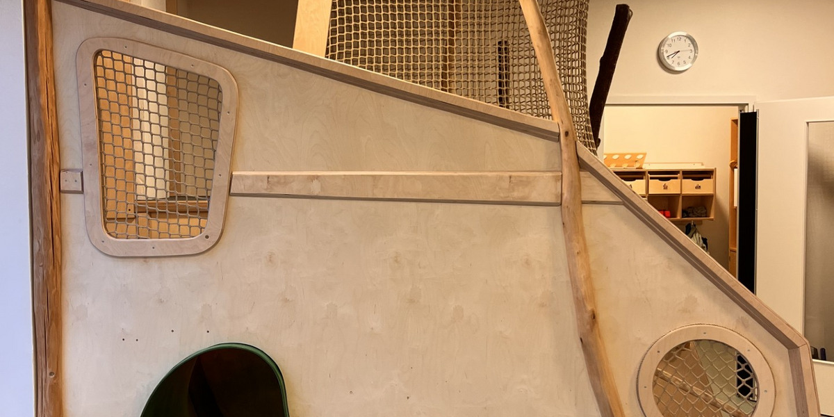 In einem hellen Raum steht eine Spielstruktur aus Holz. Sie besteht aus einer gewundenen Rutsche, einem Tunnel mit einer organischen Form und netzartigen Elementen, die zum Klettern einladen. Die Struktur ist naturnah gestaltet.