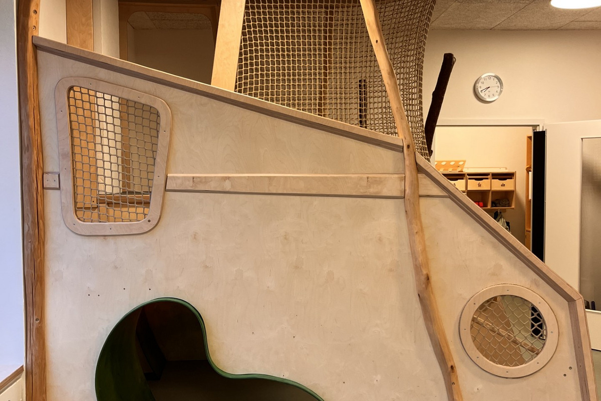In einem hellen Raum steht eine Spielstruktur aus Holz. Sie besteht aus einer gewundenen Rutsche, einem Tunnel mit einer organischen Form und netzartigen Elementen, die zum Klettern einladen. Die Struktur ist naturnah gestaltet.