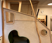 In einem hellen Raum steht eine Spielstruktur aus Holz. Sie besteht aus einer gewundenen Rutsche, einem Tunnel mit einer organischen Form und netzartigen Elementen, die zum Klettern einladen. Die Struktur ist naturnah gestaltet.