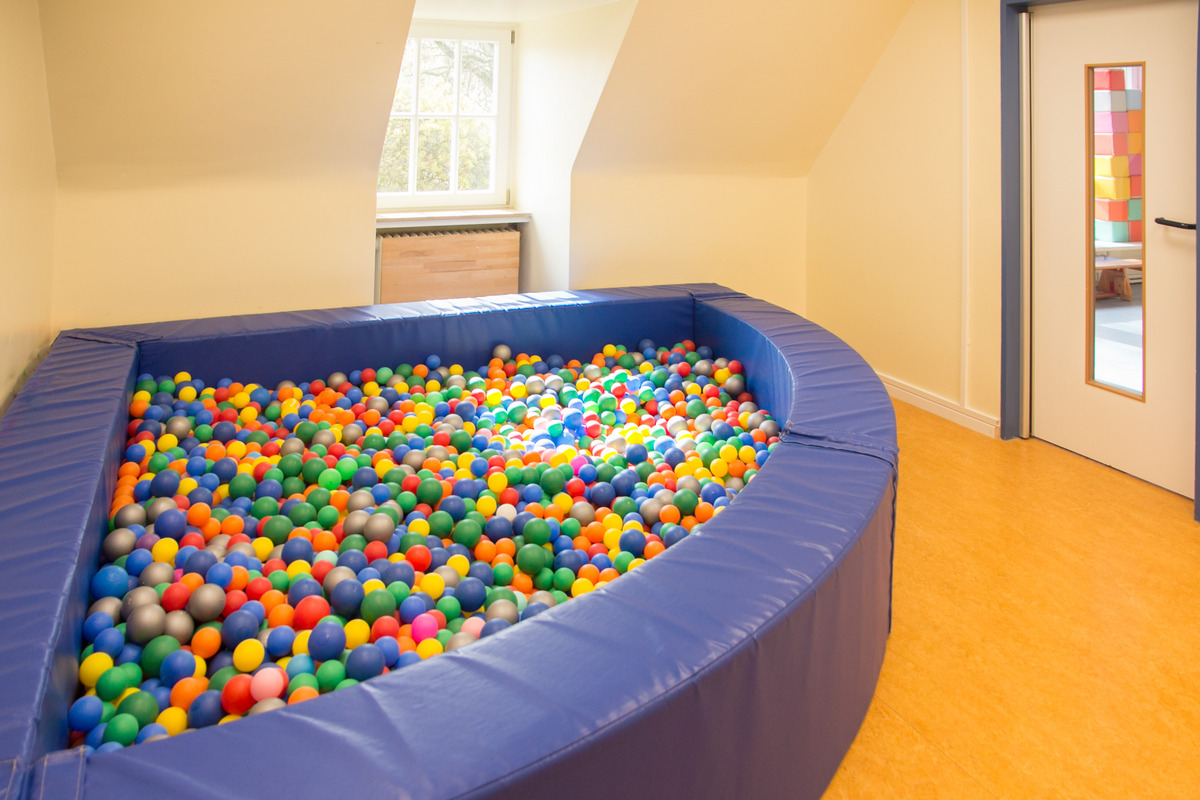 Ein großer, ovaler Bällepool aus blauen Polstern, gefüllt mit bunten Kunststoffbällen in verschiedenen Farben wie Grün, Gelb, Blau und Orange. Der Pool steht in einem hellen Raum mit einem Fenster und einer Tür im Hintergrund.