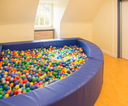 Ein großer, ovaler Bällepool aus blauen Polstern, gefüllt mit bunten Kunststoffbällen in verschiedenen Farben wie Grün, Gelb, Blau und Orange. Der Pool steht in einem hellen Raum mit einem Fenster und einer Tür im Hintergrund.