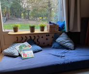 Ein gemütlicher Lese- oder Entspannungsbereich mit einem blauen Kissen und einer blauen Matte. Auf einem Tisch liegen Bücher. Im Hintergrund ist ein Fenster mit Blick auf einen Garten.
