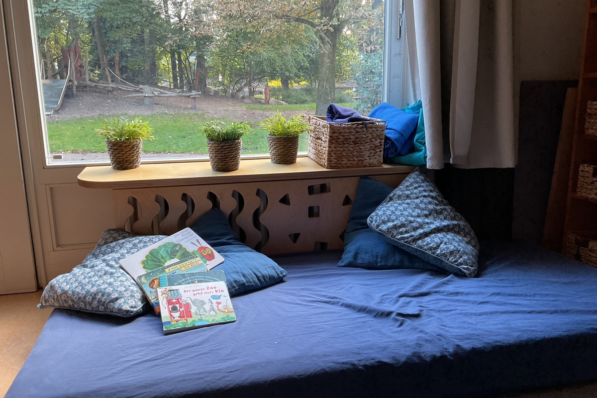 Ein gemütlicher Lese- oder Entspannungsbereich mit einem blauen Kissen und einer blauen Matte. Auf einem Tisch liegen Bücher. Im Hintergrund ist ein Fenster mit Blick auf einen Garten.