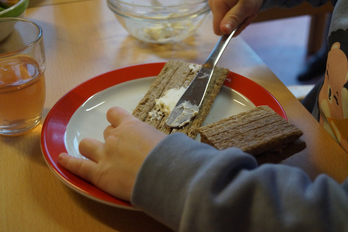 Ein Kind verteilt Butter auf einer Scheibe Brot auf einem Teller. 