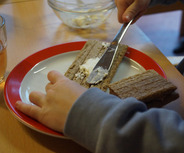 Ein Kind verteilt Butter auf einer Scheibe Brot auf einem Teller. 