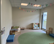 Ein heller Raum mit einem runden, grünen Teppich. An einer Wand befinden sich Spielmaterialien und eine Regale mit Büchern. Bunte Wimpel hängen an der Decke. Es gibt eine große Fensterfront, die einen Blick nach draußen ermöglicht. Ein Stuhl und einige blaue Matten sind ebenfalls sichtbar.