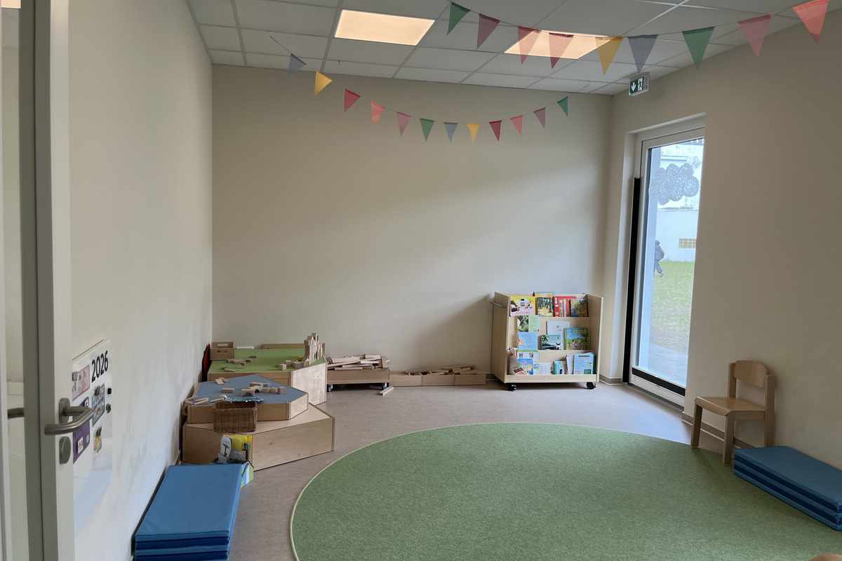 Ein heller Raum mit einem runden, grünen Teppich. An einer Wand befinden sich Spielmaterialien und eine Regale mit Büchern. Bunte Wimpel hängen an der Decke. Es gibt eine große Fensterfront, die einen Blick nach draußen ermöglicht. Ein Stuhl und einige blaue Matten sind ebenfalls sichtbar.