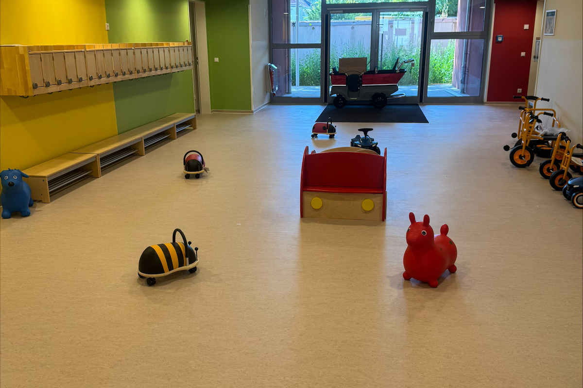 Ein heller Flur mit bunten Wänden, ausgestattet mit Spielzeugen wie kleinen Fahrzeugen und Plüschtieren. Im Hintergrund sind große Fenster, die auf einen Garten blicken, und es gibt eine Sitzecke mit einem roten Sofa.