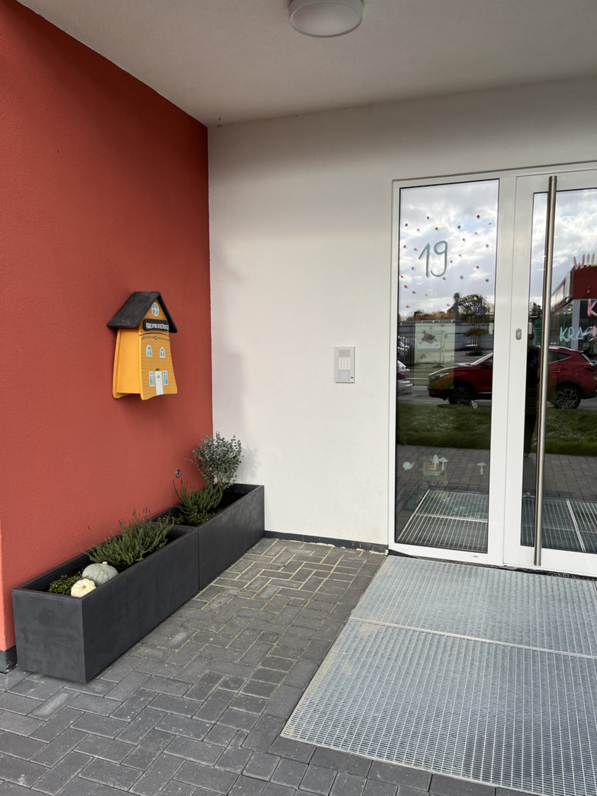 Der Eingangsbereich eines Gebäudes mit einer rot-weißen Wand. An der Wand hängt ein Briefkasten in Form eines Hauses. 