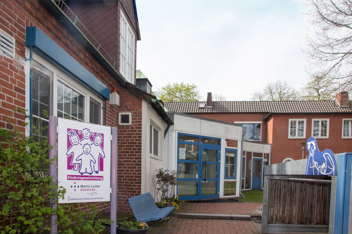 Das Bild zeigt den Eingangsbereich einer Kindertagesstätte mit einem bunten Schild, das die Aufschrift "Kindertageseinrichtung Martin Luther Gemeinde" trägt. Im Hintergrund sind die Gebäude der Einrichtung und eine kleine Bank zu sehen.
