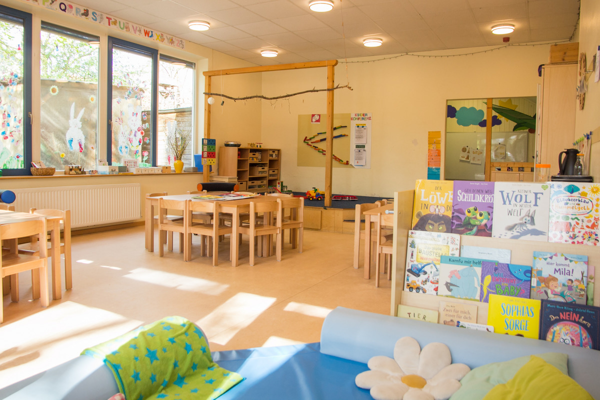 Ein heller Gruppenraum mit Holzmöbeln ausgestattet. In der Mitte stehen Tische und Stühle, an der Wand sind Bücherregale mit verschiedenen Kinderbüchern. Große Fenster lassen viel Licht herein. An den Wänden sind bunte Malereien und Dekorationen sichtbar.
