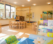 Ein heller Gruppenraum mit Holzmöbeln ausgestattet. In der Mitte stehen Tische und Stühle, an der Wand sind Bücherregale mit verschiedenen Kinderbüchern. Große Fenster lassen viel Licht herein. An den Wänden sind bunte Malereien und Dekorationen sichtbar.