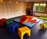 Ein Spielzimmer mit bunten, weichen Kissen und Schaumstoffelementen ist zu sehen. Es gibt verschiedene farbige Sitzwürfel in Rot, Grün, Blau und Gelb, sowie eine Kletterstruktur mit Rutschen in Orange und Blau. Eine Tunnelröhre in Grün ergänzt die Spielmöglichkeiten.