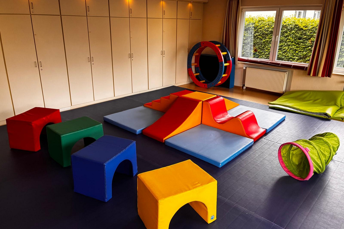 Ein Spielzimmer mit bunten, weichen Kissen und Schaumstoffelementen ist zu sehen. Es gibt verschiedene farbige Sitzwürfel in Rot, Grün, Blau und Gelb, sowie eine Kletterstruktur mit Rutschen in Orange und Blau. Eine Tunnelröhre in Grün ergänzt die Spielmöglichkeiten.