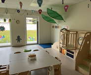 Ein heller Raum. An einem Tisch stehen Stühle, im Hintergrund gibt es eine Kletteranlage aus Holz. Durch große Fenster ist der Blick ins Freie möglich, mit bunten Dekorationen an den Fenstern.