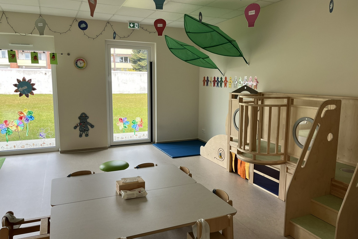 Ein heller Raum. An einem Tisch stehen Stühle, im Hintergrund gibt es eine Kletteranlage aus Holz. Durch große Fenster ist der Blick ins Freie möglich, mit bunten Dekorationen an den Fenstern.