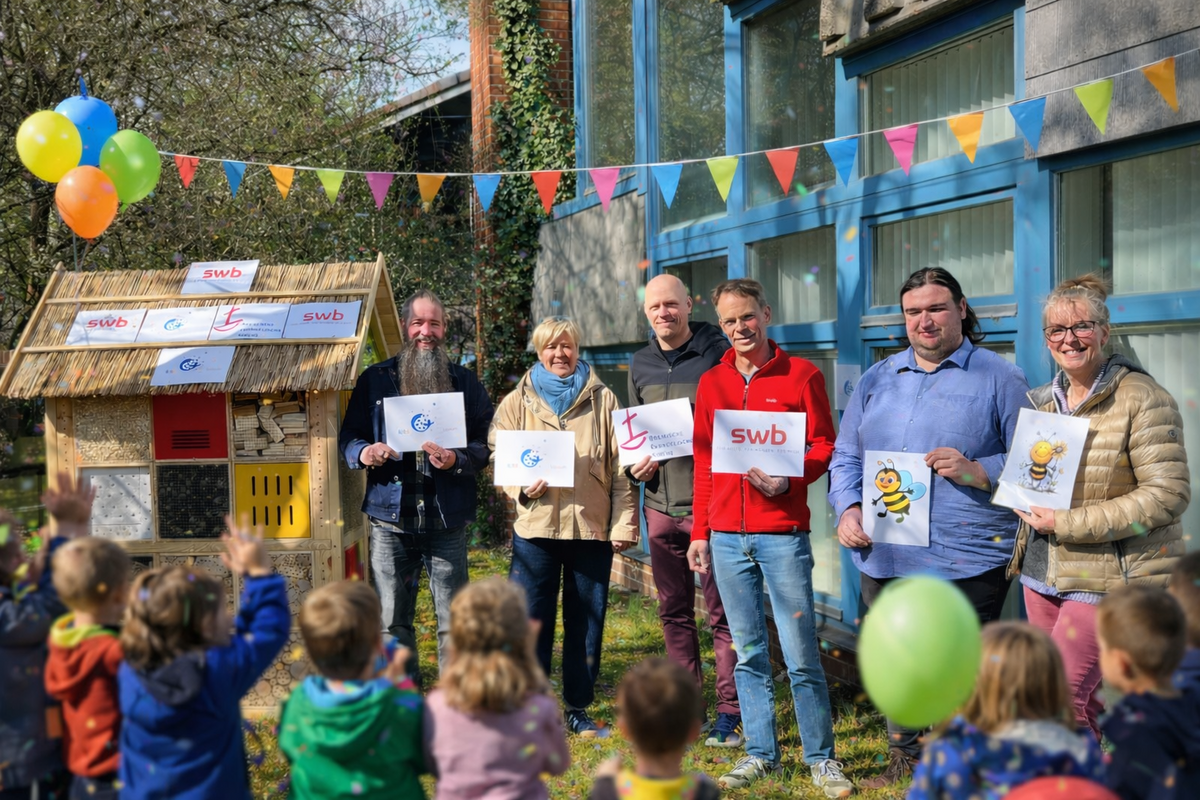 Eine Gruppe von sechs Erwachsenen steht vor einem kleinen, bunten Insektenhotel, während Kinder im Vordergrund applaudieren. Jeder Erwachsene hält ein Plakat mit einem Logo oder einem Bienenmotiv in der Hand. Im Hintergrund sind Luftballons und Wimpel sichtbar, die eine festliche Atmosphäre schaffen.
