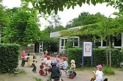 Eine Gruppe von Kindern fährt fröhlich auf Dreirädern in einem Gartenbereich. Um sie herum sind Bäume und Sträucher. 