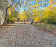 Ein herbstlicher Außenbereich mit Laub auf dem Boden. Auf einem Weg sind mehrere Kinder zu sehen, die in Richtung eines Spielplatzes gehen, umgeben von bunten, herbstlichen Bäumen im Hintergrund. 