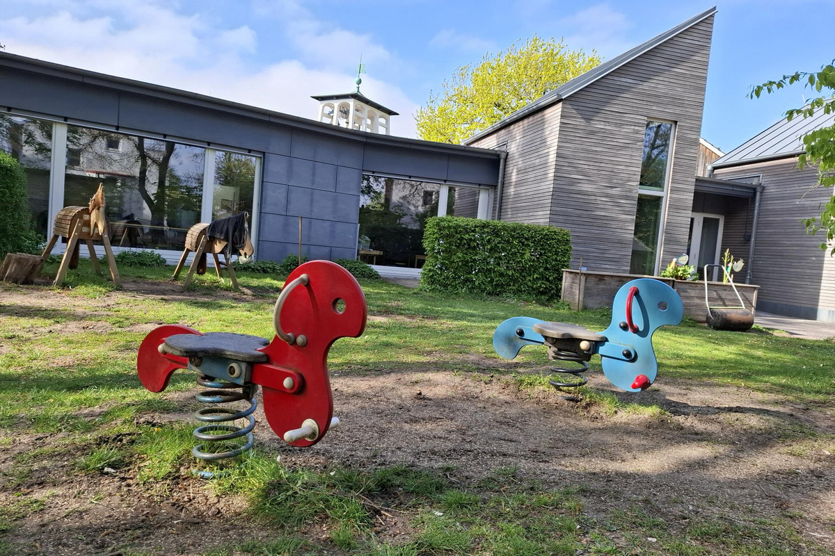 Auf einem Spielplatz sind zwei Federwippe in Form von Pferden, eine rot und eine blau. Im Hintergrund steht ein modernes Gebäude mit großen Fenstern und einer grünen Hecke. Der Rasen ist leicht uneben und es ist sonnig.