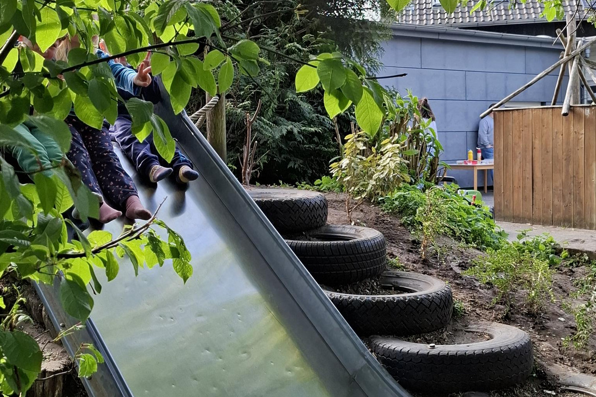 Eine metallene Rutsche in einem Garten, umgeben von grünen Blättern und Pflanzen. Im Hintergrund sind Kinder zu sehen, die sich in der Nähe der Rutsche befinden. Reifenstapel dienen als Treppe zur Rutsche.