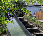 Eine metallene Rutsche in einem Garten, umgeben von grünen Blättern und Pflanzen. Im Hintergrund sind Kinder zu sehen, die sich in der Nähe der Rutsche befinden. Reifenstapel dienen als Treppe zur Rutsche.