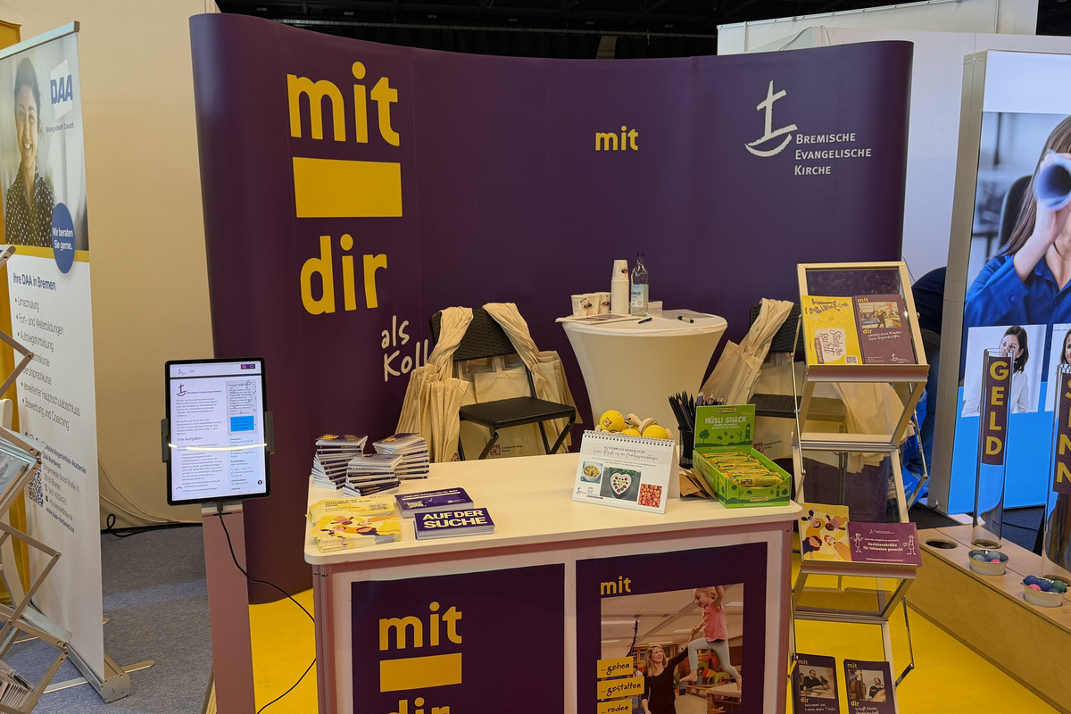 Der Messestand der Evangelischen Kirche zeigt eine lila Wand mit dem Schriftzug "mit dir als Kollegin", einen Tisch mit verschiedenen Informationsmaterialien und Broschüren, sowie Ständern mit Flyern. 