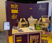 Der Messestand der Evangelischen Kirche zeigt eine lila Wand mit dem Schriftzug "mit dir als Kollegin", einen Tisch mit verschiedenen Informationsmaterialien und Broschüren, sowie Ständern mit Flyern. 