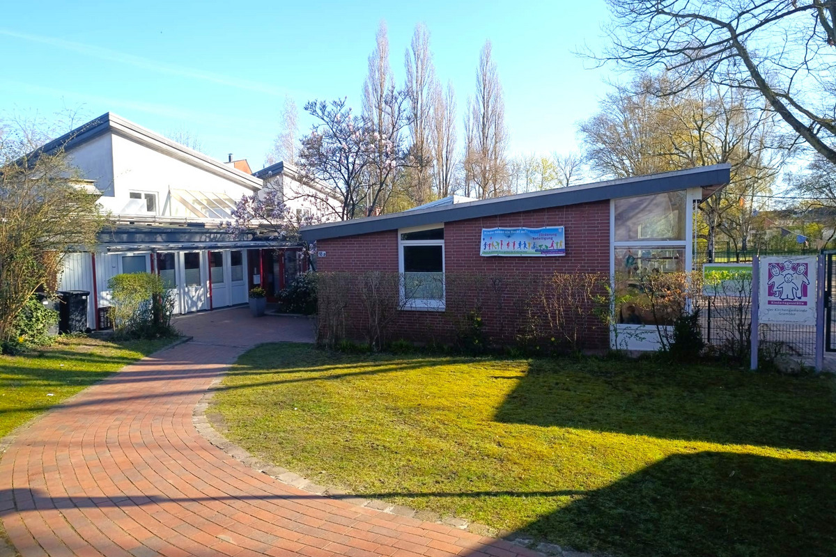 Ein gepflasterter Weg führt zu einem niedrigen Gebäude mit großen Fenstern und einem modernen Dach. Vor dem Gebäude sind Sträucher und ein kleiner Blumenkübel sichtbar. Im Hintergrund sind Bäume und ein blauer Himmel zu sehen.