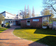 Ein gepflasterter Weg führt zu einem niedrigen Gebäude mit großen Fenstern und einem modernen Dach. Vor dem Gebäude sind Sträucher und ein kleiner Blumenkübel sichtbar. Im Hintergrund sind Bäume und ein blauer Himmel zu sehen.