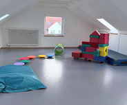 Ein heller Raum mit schrägen Decken. Auf dem Boden liegen bunte Kissen, ein grüner Tunnel und ein Kletterblock aus farbigen Schaumstoffelementen. In der Ecke befindet sich eine große, weich gepolsterte Matte. Fenster an der Wand lassen viel Tageslicht herein.