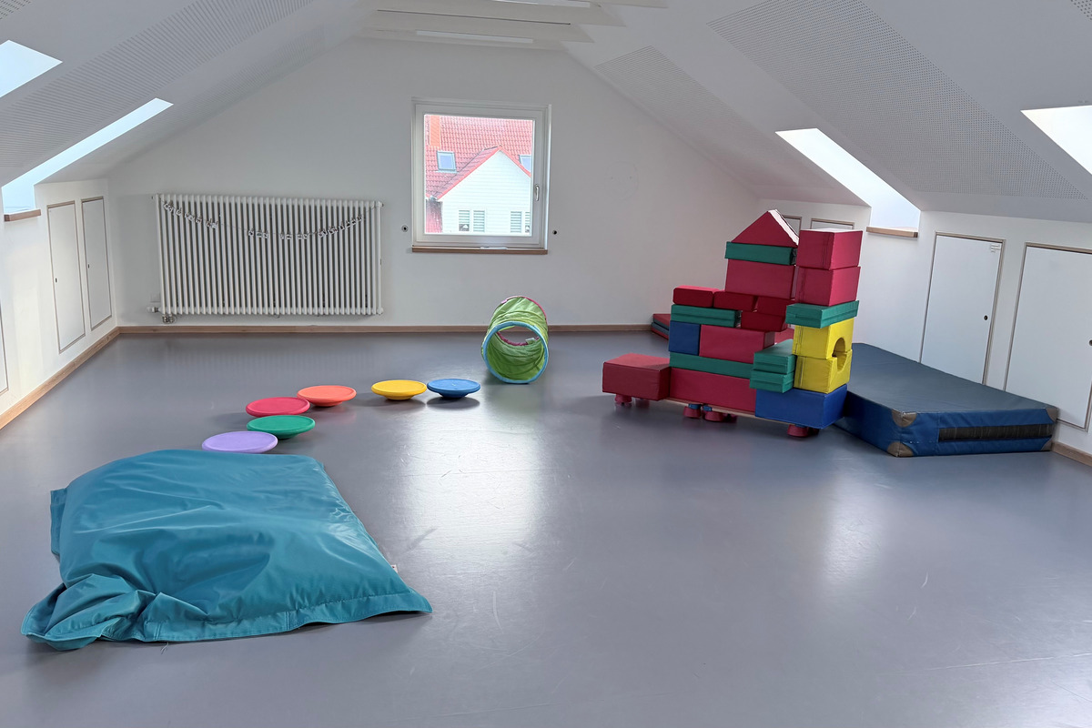 Ein heller Raum mit schrägen Decken. Auf dem Boden liegen bunte Kissen, ein grüner Tunnel und ein Kletterblock aus farbigen Schaumstoffelementen. In der Ecke befindet sich eine große, weich gepolsterte Matte. Fenster an der Wand lassen viel Tageslicht herein.